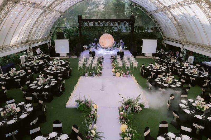 Không gian tổ chức sự kiện được bố trí với bàn ghế trải đều, lối đi trang trí hoa và hệ thống ánh sáng sân khấu hiện đại