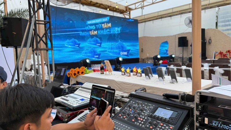 Kỹ thuật viên đang điều khiển thiết bị âm thanh ánh sáng trong sự kiện ngoài trời với màn hình LED và bàn ghế sắp xếp chuẩn bị.