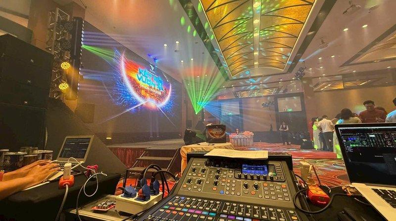 Bàn điều khiển âm thanh và ánh sáng được bố trí trong một hội trường sự kiện với hệ thống đèn màu sắc rực rỡ và màn hình lớn phía sau sân khấu